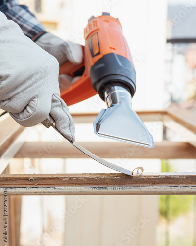 Adult carpenter wearing protective leather gloves, using a heat gun and a wood scraper to remove paint from an old wooden window. Construction industry, carpentry. Restoration. 
