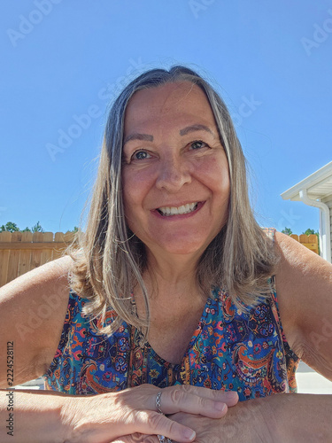 Mature woman, silver-gray hair, blue eyes, broad smile, natural beauty, authentic, confident, joyful, friendly, outdoor portrait, bright blue sky, sunlight, colorful patterned top, sleeveless blouse, 