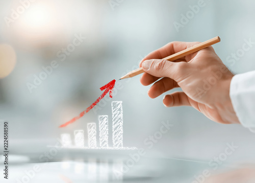 A hand holding a pencil draws a red upward arrow on a chalk style bar chart, symbolizing growth and success in business or finance.