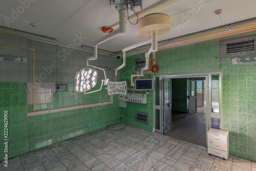 An empty operating room with green tiled walls contains ceiling-mounted surgical lights, a monitor and an open doorway to a corridor