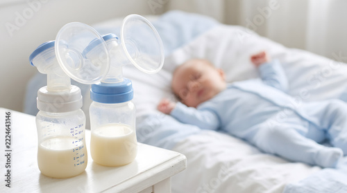 Double electric breast pump with bottle of milk on white table beside sleeping newborn baby