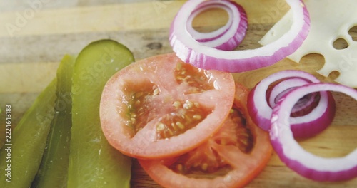 Displaying stack of sliced red tomatoes on wooden board, with pickles, onion rings, Swiss cheese