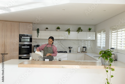 Asian woman wearing apron pouring measured ingredient from jug into bowl on kitchen island