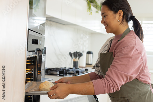 Asian woman placing round dough on baking tray into built-in oven in home kitchen, copy space