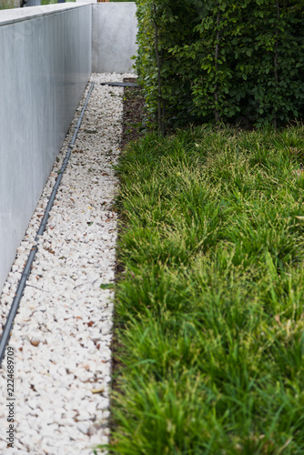 A decorative gravel path runs alongside a modern wall and a dense green hedge.