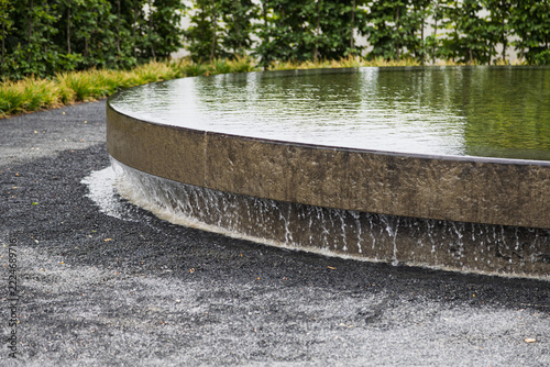 Water cascades over the curved edge of a circular stone fountain into a gravel-covered ground.