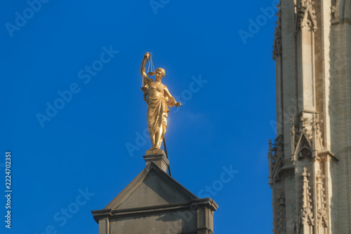 A clear Antwerp rooftop moment with a golden Lady Justice standing above dark stone, holding scales aloft and a sword lowered, her bright silhouette set sharply against the blue sky.