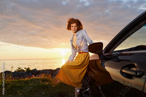 A stylish woman steps from a car at sunset, wearing a yellow skirt and denim jacket, capturing a spontaneous outdoors lifestyle moment with confidence.