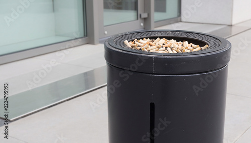 Outdoor Cigarette Butt Disposal Bin Filled with Used Cigarette Filters Near a Modern Commercial Building