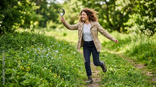Curious woman exploring nature with a magnifying glass walking on a path through lush greenery and wildflowers