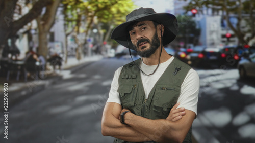 Obraz na plátně Man with beard wearing fishing vest and wide hat, arms crossed facing camera on sunlit urban street near cars and trees; quiet determination