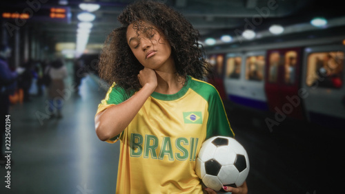 Fotografie Woman wearing brasil jersey holding football with hand on neck in building platform; tiredness resilience