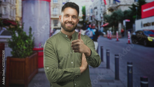 Man with beard in green shirt smiling with arms crossed and occasional thumbs up gesture, bare right hand visible, standing on city street with bollards and planter; cheerful confidence.