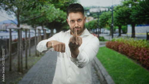 Obraz na plátně Man with beard showing a forward fist and beckoning hand on street promenade by trees and water, looking at camera; assertive confidence