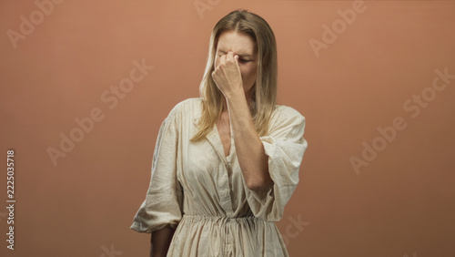 Obraz na plátně Blonde woman pinches nose bridge between eyes and grimaces against peach studio backdrop; disgust discomfort avoidance