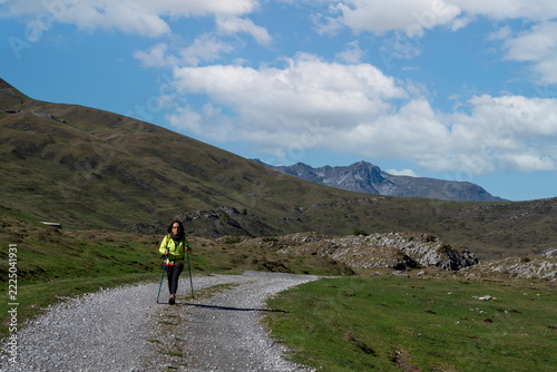 female hiker wearing a yellow jacket and carrying trekking poles walks along a gravel path through green mountain landscapes with rocky peaks in the background under a blue cloudy sky.