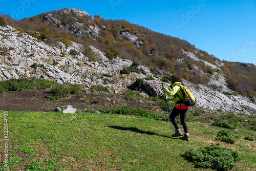 side view of a woman wearing a bright yellow jacket, carrying a backpack and trekking poles, hiking up a grassy mountain hill 