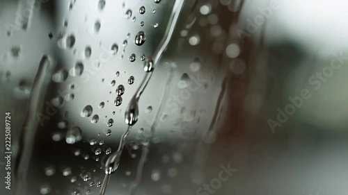 Slow motion static tilt pan of raindrops sliding down glass window pane with blurred outdoor background