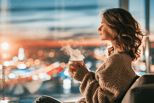 A serene young woman with wavy hair relaxes in a cozy sweater, holding a steaming cup of coffee while enjoying a sunset view over a harbor with city lights.