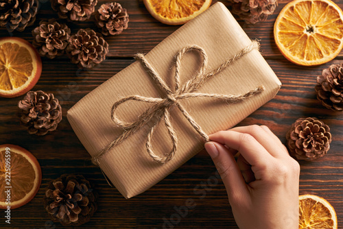 A woman's hand reaching for a gift wrapped in brown kraft paper and tied with twine, surrounded by dried orange slices and pine cones on a rustic wooden background.