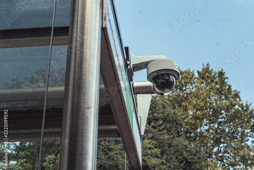 A security camera is mounted on the corner of a modern building, overlooking a leafy area. The camera provides surveillance and security in an urban environment.