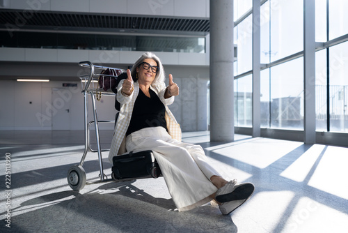 White woman traveler showing thumbs up sitting on luggage trolley