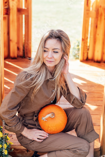 Woman holding a pumpkin at an outdoor autumn farm market surrounded by wooden shelves filled with pumpkins, warm sunlight, and seasonal harvest decorations