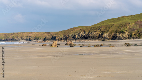 A broad sandy beach with scattered rocks and exposed tidal flats beneath green coastal hills and rocky cliffs near Porth Colmon on the Welsh coast. Calm, natural coastal landscape in soft daylight wit
