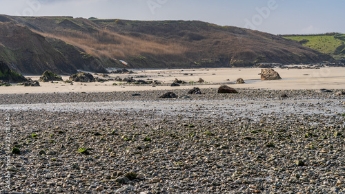A rugged pebble beach with scattered rocks and shallow tidal water beneath eroded coastal cliffs near Porth Colmon on the Welsh coast. Natural seaside landscape in clear daylight with textured stones 