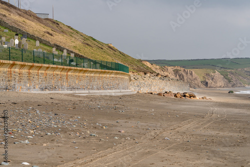 A quiet sandy beach with a long stone seawall, pebble-strewn shore, and rugged coastal cliffs under soft daylight at Aberdaron on the Llŷn Peninsula. Natural coastal scene with open space and gentle w