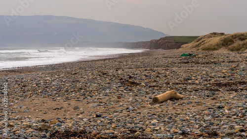 A wide pebble beach with scattered driftwood, rolling waves, and distant coastal cliffs under soft, hazy light near Llanengan on the Llŷn Peninsula. Natural coastal scene with strong textures and an u