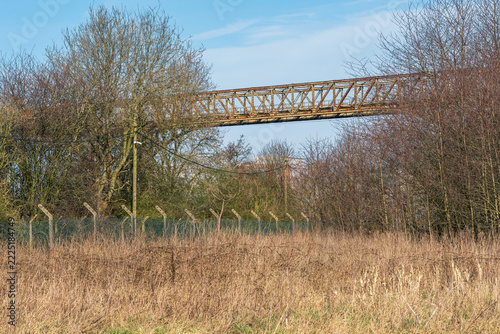 Warrington Transporter Bridge, Chesire, England, UK: Rusty steel industrial bridge stretches above dry grass and leafless trees behind a security fence. A quiet scene symbolizing industrial decay and 