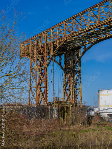 Warrington Transporter Bridge, Chesire, England, UK: Rusty steel industrial bridge rises above trees with modern factory buildings in the background. A contrast between industrial heritage and contemp