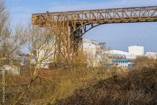 Warrington Transporter Bridge, Chesire, England, UK: Rusty steel industrial bridge rises above trees with modern factory buildings in the background. A contrast between industrial heritage and contemp