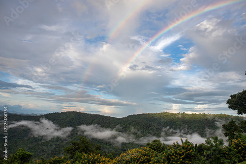 Double rainbow over misty green mountains.A spectacular double rainbow arcs across a cloudy sky over a scenic mountain valley covered in morning mist and yellow wildflowers.