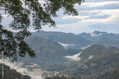 Mountain valley with morning mist under tree branch.A high-angle view of a tropical mountain valley with white fog and buildings in the distance, framed by tree leaves in Khao Yai, Thailand.