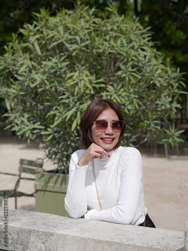Asian woman wearing sunglasses smiling while resting her chin on her hand against a stone ledge in a park