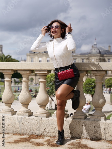Asian woman in sunglasses smiling and adjusting her hair while leaning against a stone balustrade in Paris