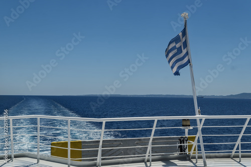 Greek flag fluttering on the stern railing of a ferry creating a foamy wake across the blue sea in Rhodes, Greece.
