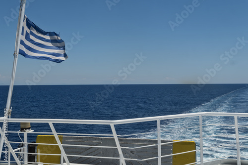 Greek flag fluttering on the stern railing of a ferry creating a foamy wake across the blue sea in Rhodes, Greece.