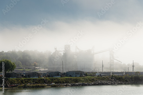 Industrial grain storage silos and conveyor structures partially obscured by dense fog near a waterfront residential area. Human impact conception.