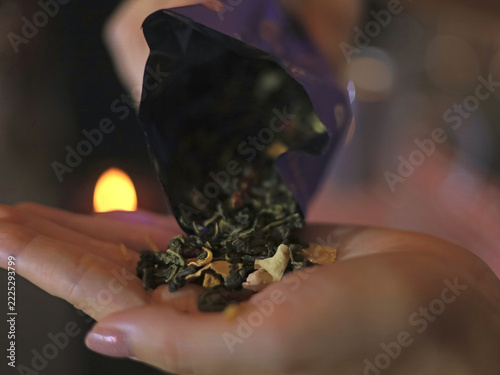 Fotografie Loose leaf tea with dried fruit pieces pours from a dark pouch into an open palm against soft candlelight