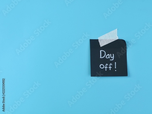 Black adhesive note with day off text handwritten attached with masking tape on blue background with copy space. Perfect concept for vacation, holiday, time off, work life balance, and office leave