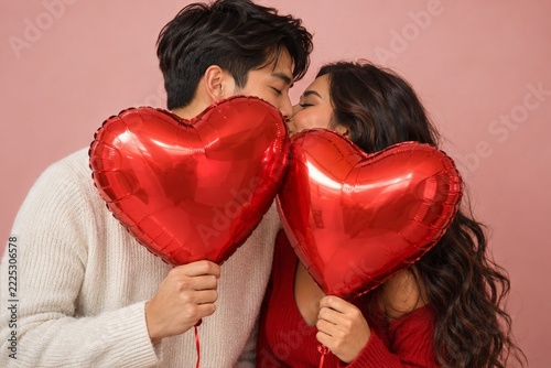 Young couple sharing a tender kiss behind glossy red heart shaped balloons