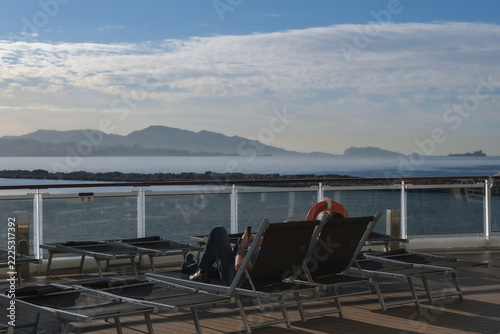 Pooldeck or swimmingpool with sun deck on modern cruiseship cruise ship liner in port with sun loungers ready for cruising to Mediterranean on sunny day with city skyline in background