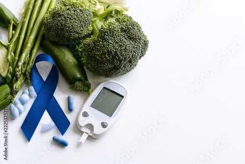 Diabetes awareness concept featuring blue ribbon and blood glucose monitor with fresh green vegetables on white background for health.