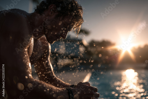 Exhausted male swimmer resting by pool edge during sunset with glowing water droplets