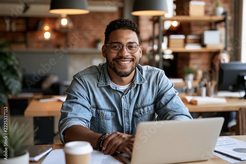 Smiling young businessman sitting at desk with laptop in modern open space office with brick wall