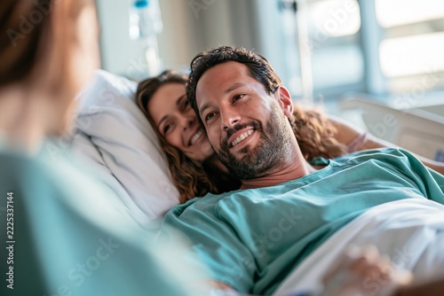 Smiling male patient resting in hospital bed with supportive partner visiting in recovery room