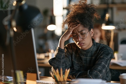 Stressed young woman holding her head at office desk working late night with dim desk lamp